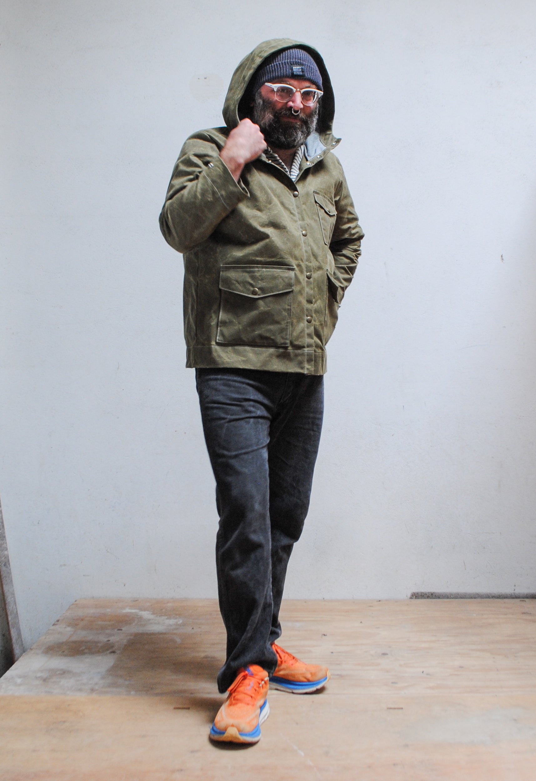 Person wearing a green jacket with a hood, black pants, and orange shoes on a wooden floor.