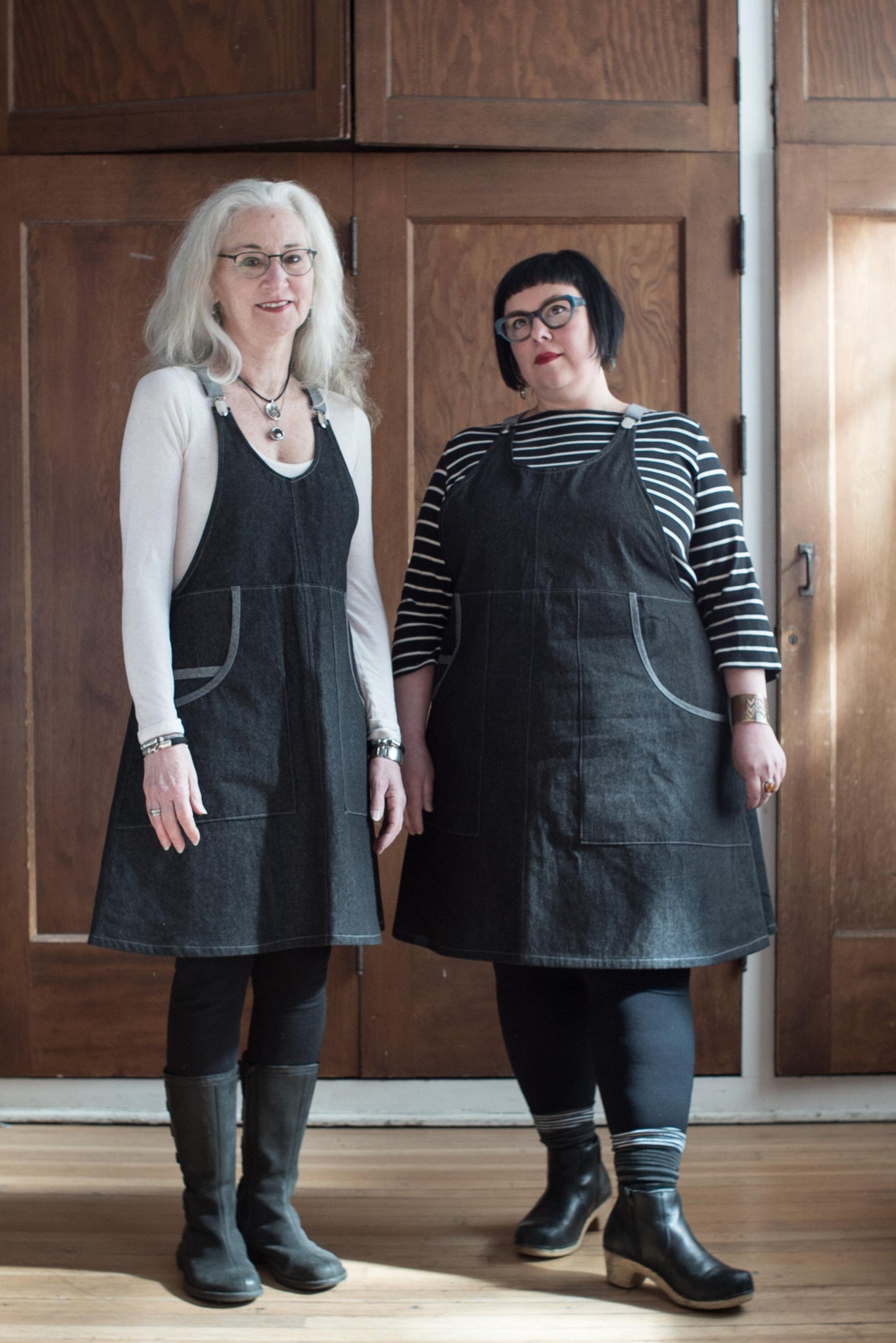3/4 angle view of two models wearing black denim suspender dress. 100% cotton 8oz denim has a black wash and reads as very dark grey. The fabric is sturdy, hardworking yet lightweight. The pockets feature a contrasting self-trim. 