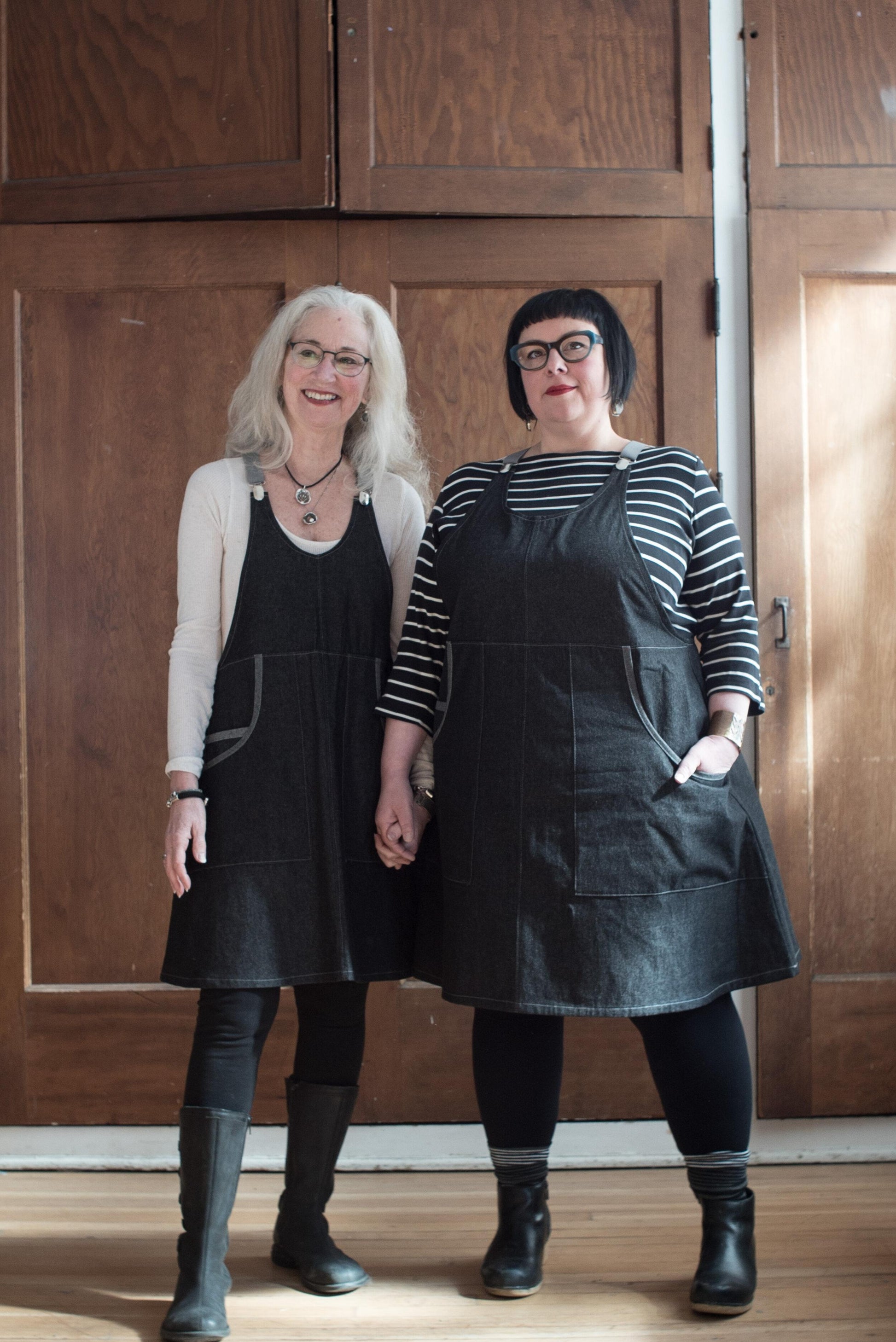 Front view of two models wearing black denim suspender dress.100% cotton 8oz denim has a black wash and reads as very dark grey. The fabric is sturdy, hardworking yet lightweight. The pockets feature a contrasting self-trim. 