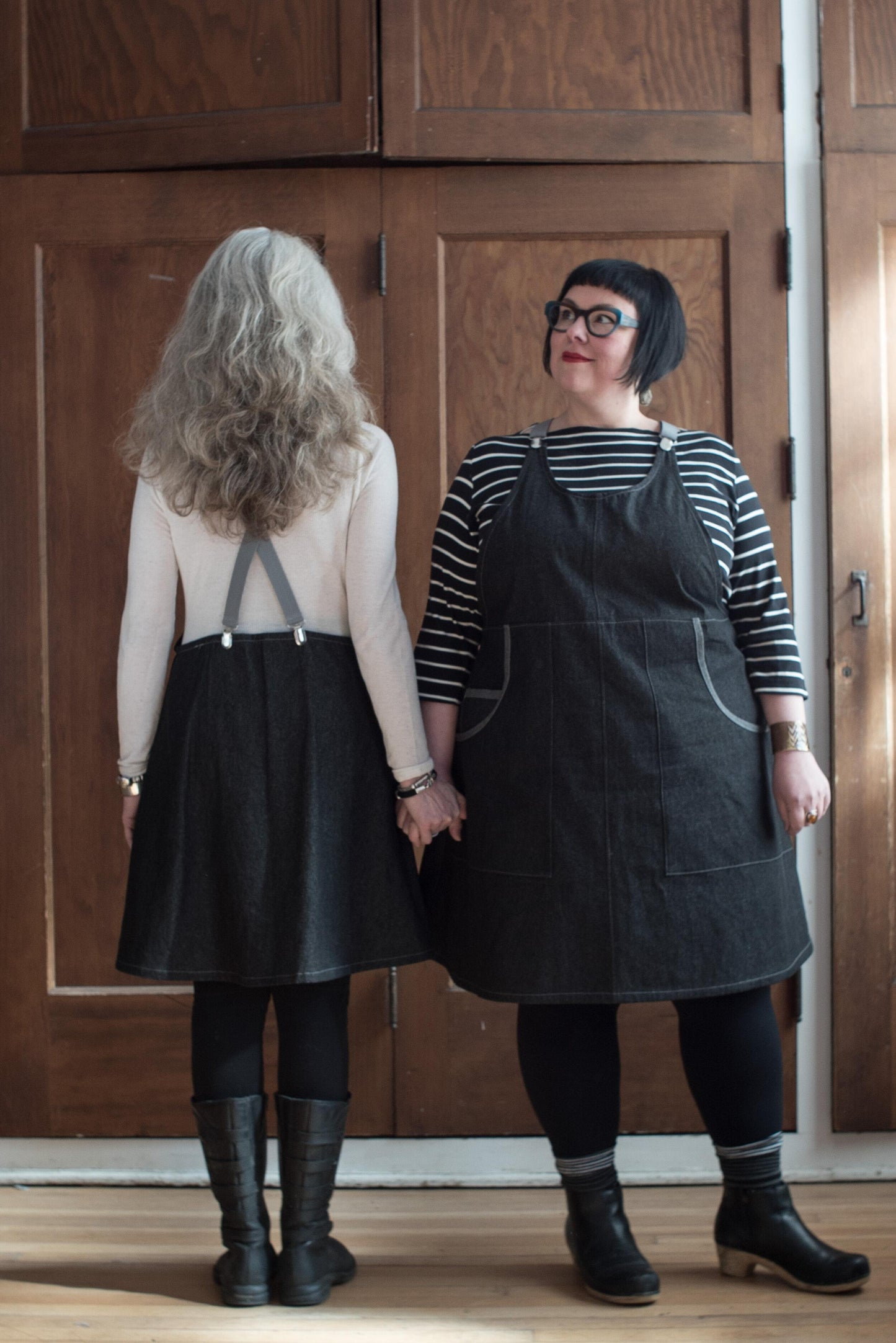 Front and back view of black denim suspender dress on two models. 100% cotton 8oz denim has a black wash and reads as very dark grey. The fabric is sturdy, hardworking yet lightweight. The pockets feature a contrasting self-trim. 